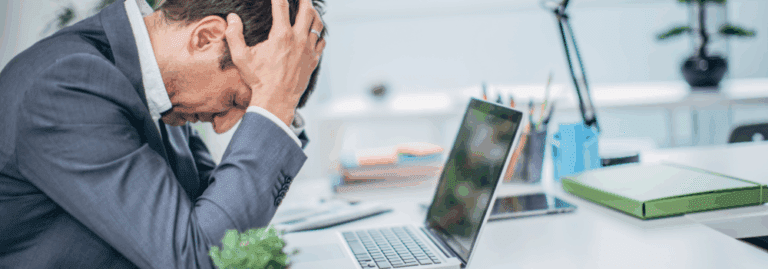 Man sitting at desk holding his head in stress, representing burnout and staffing challenges in mental health services.